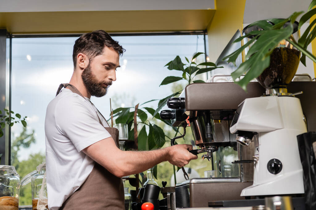 bearded barista in apron holding holder near coffee machine while working in coffee shop - Photo, Image