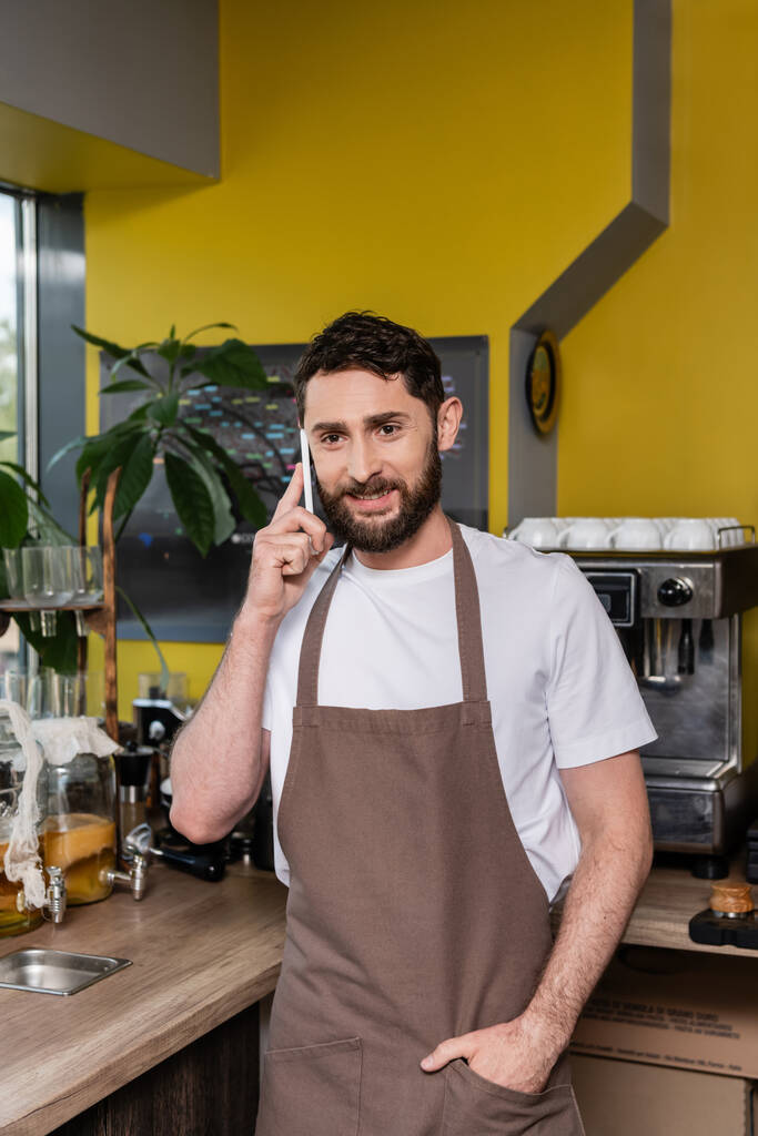 usmívající se vousatý barista v zástěře mluvit na smartphone při práci v kavárně na pozadí - Fotografie, Obrázek