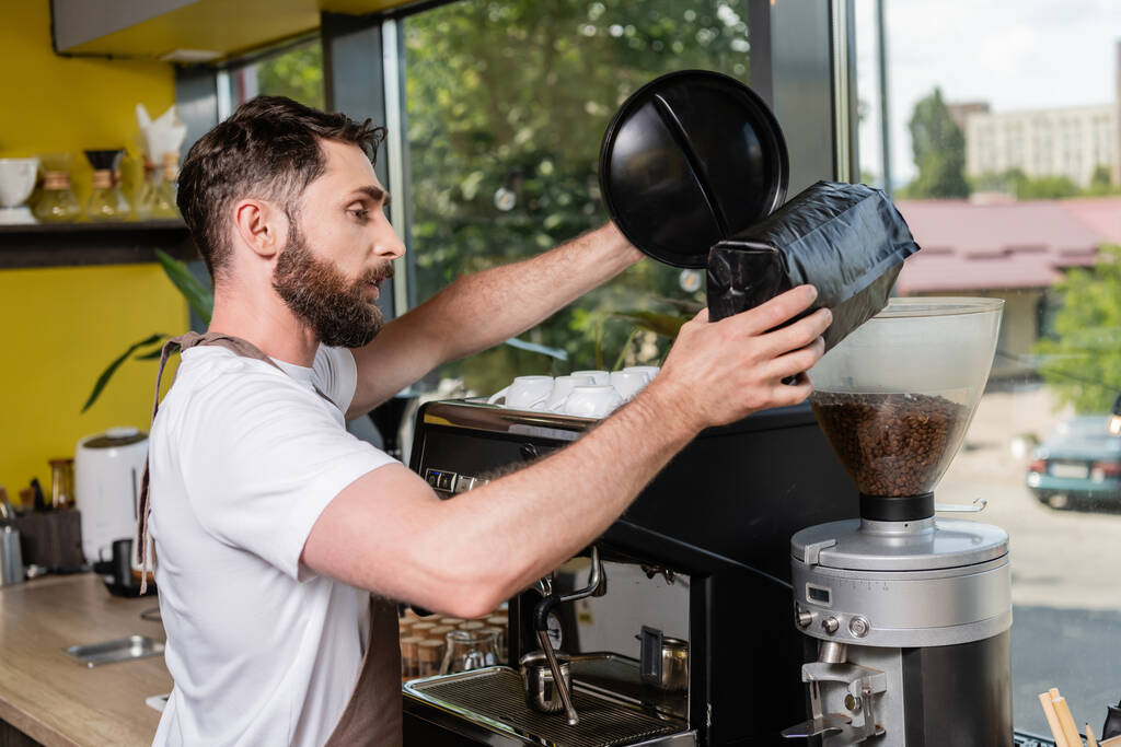 side view of barista in apron pouring coffee beans in grinder while working in coffee shop - Photo, Image