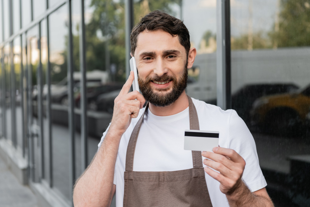 fröhlicher Barista hält Kreditkarte in der Hand und unterhält sich auf der Straße im Hintergrund mit dem Smartphone - Foto, Bild