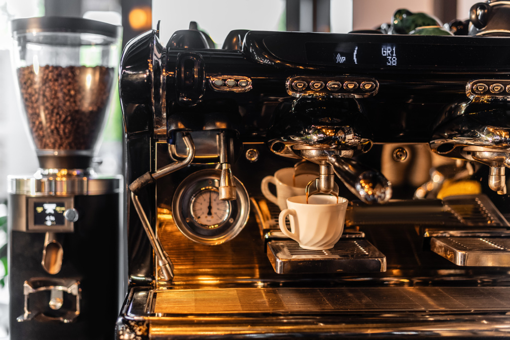 káva nalévání z kávovaru v šálku v blízkosti rozmazané mlýnek se slunečním světlem v kavárně - Fotografie, Obrázek
