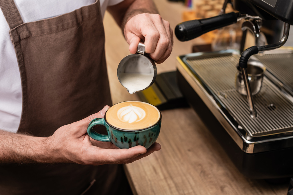 ミルクを注いでカフェのコーヒーマシンの近くでカプチーノを作るエプロンのバリスタのクロップビュー - 写真・画像