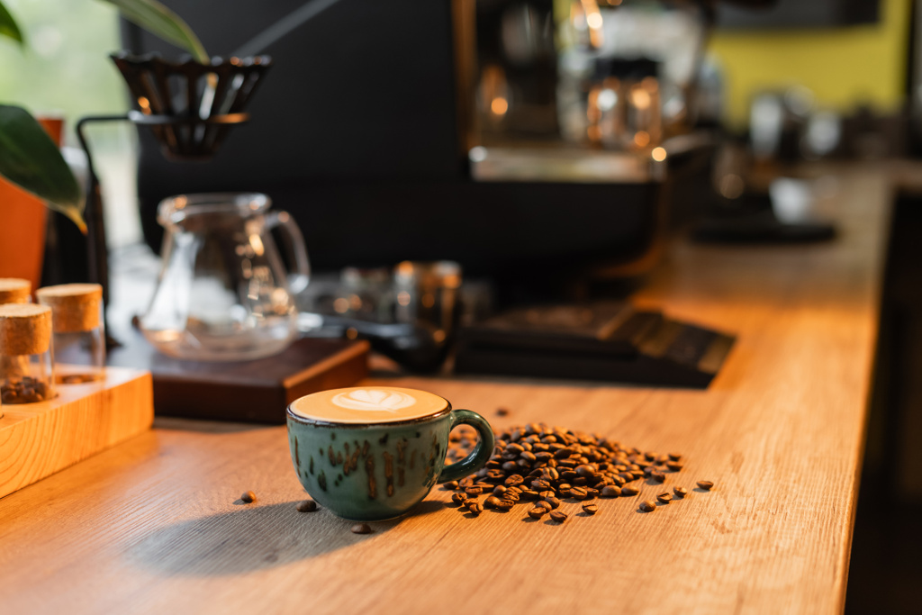 φλιτζάνι καπουτσίνο και κόκκους καφέ στον πάγκο εργασίας σε θολή καφετέρια στο παρασκήνιο - Φωτογραφία, εικόνα