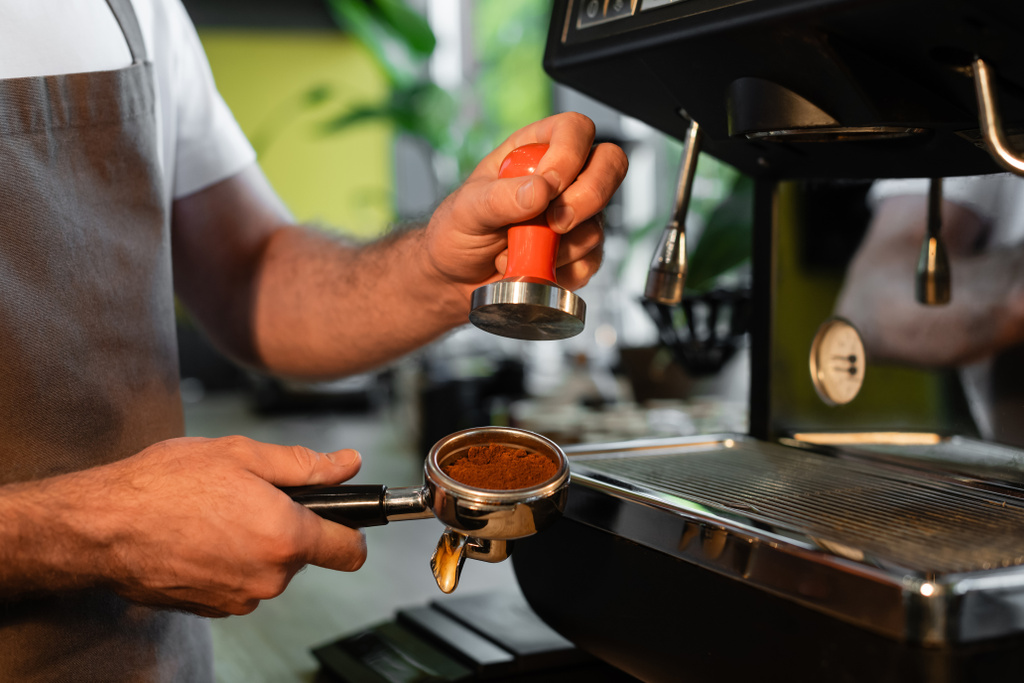 カフェのコーヒーマシン近くのホールダーにプレスとコーヒーを保持するエプロンのバリスタのクロップビュー - 写真・画像