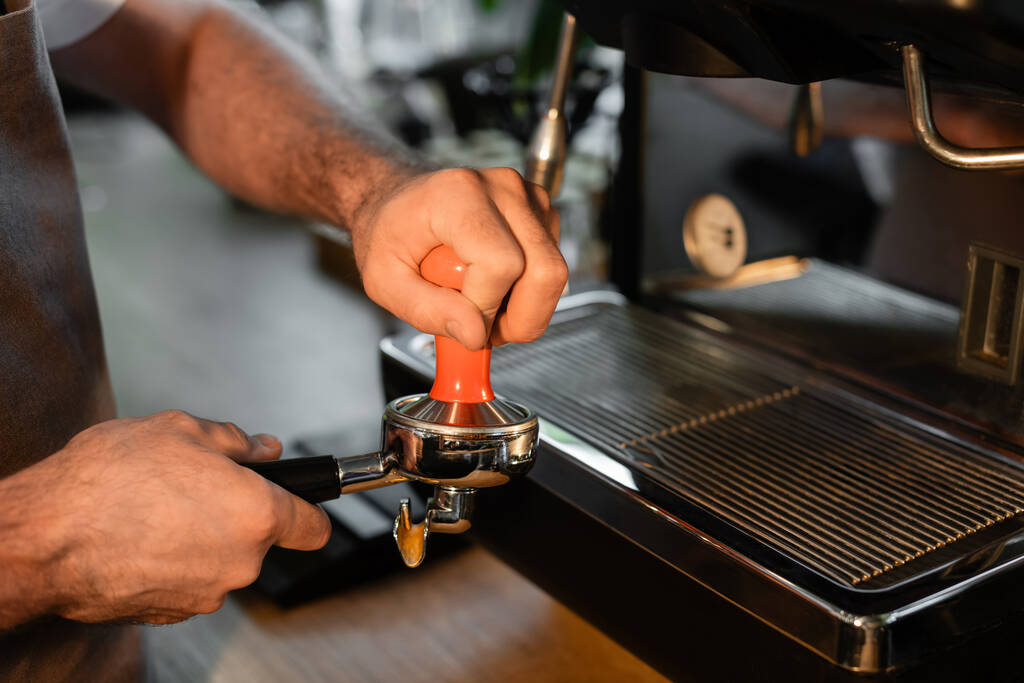 vista ritagliata di barista in grembiule che tiene il supporto della pressa vicino alla macchina del caffè offuscata in caffè - Foto, immagini