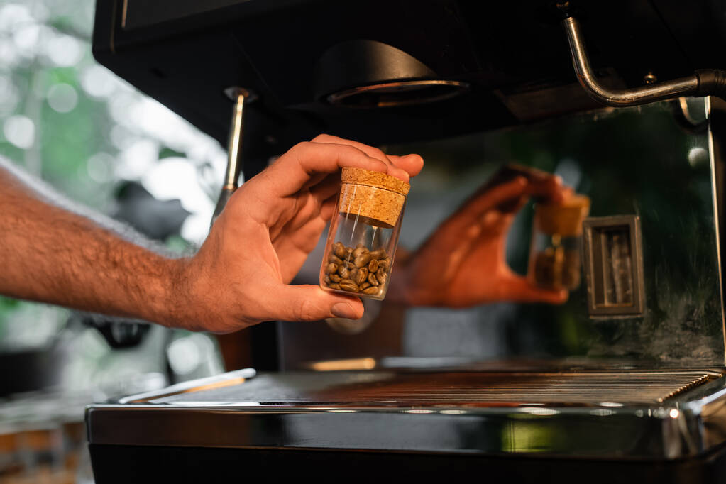 vista cortada de barista segurando grãos de café em jarra perto de máquina de café borrada no café - Foto, Imagem