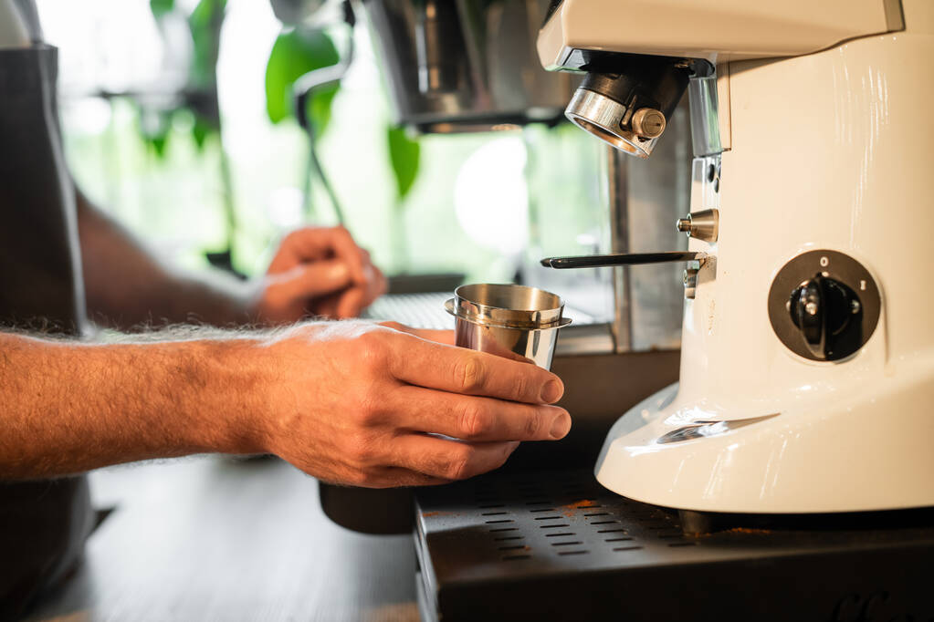 bijgesneden weergave van barista holding beker in de buurt van koffiemolen tijdens het werk in de coffeeshop - Foto, afbeelding