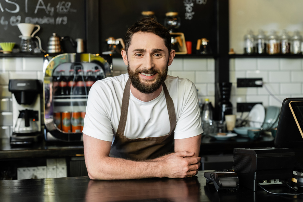 vrolijke barista met baard kijkend naar camera terwijl hij in de buurt van een bar in de coffeeshop staat - Foto, afbeelding