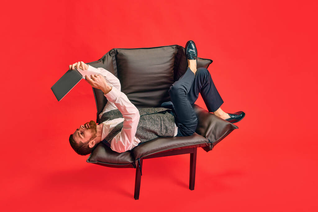 Elegante hombre de negocios acostado en un sillón, sosteniendo la sobrecarga del portátil y trabajando en una posición extraña contra el fondo rojo del estudio. Concepto de negocio, rutina de trabajo, plazos, freelance, oficina, anuncio - Foto, Imagen