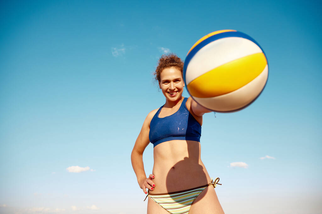 Młoda uśmiechnięta kobieta trzymająca piłkę do siatkówki, pozująca na tle błękitnego nieba. Letni trening na świeżym powietrzu. Pojęcie sportu, aktywnego i zdrowego stylu życia, hobby, lato, reklama - Zdjęcie, obraz