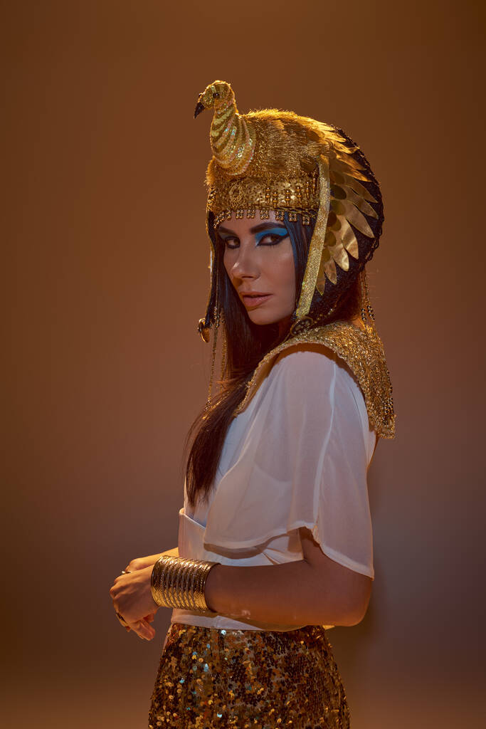 Stilvolle Frau in ägyptischer Kleidung und Kopfbedeckung blickt in die Kamera, während sie isoliert auf braun posiert - Foto, Bild
