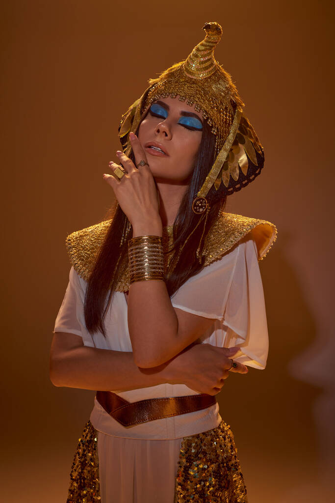 Стильная женщина с смелым макияжем и египетский наряд позирует с закрытыми глазами на коричневом фоне - Фото, изображение