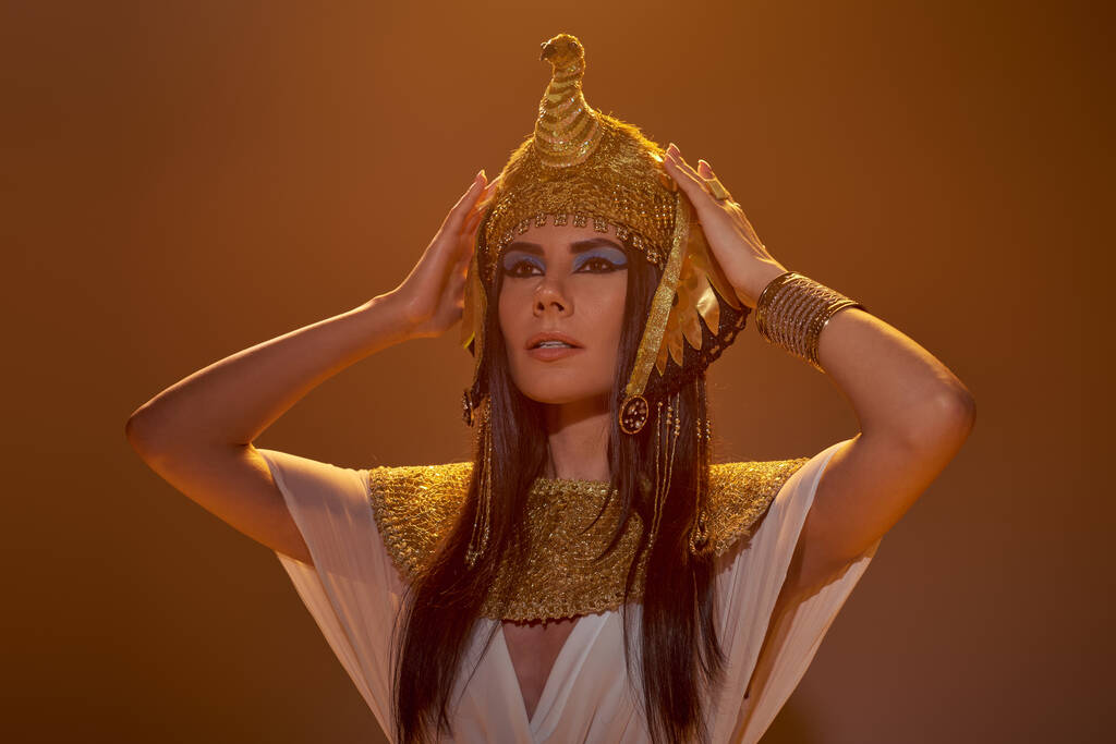 Schöne brünette Frau im ägyptischen Outfit berühren Kopfschmuck im Licht auf braunem Hintergrund - Foto, Bild