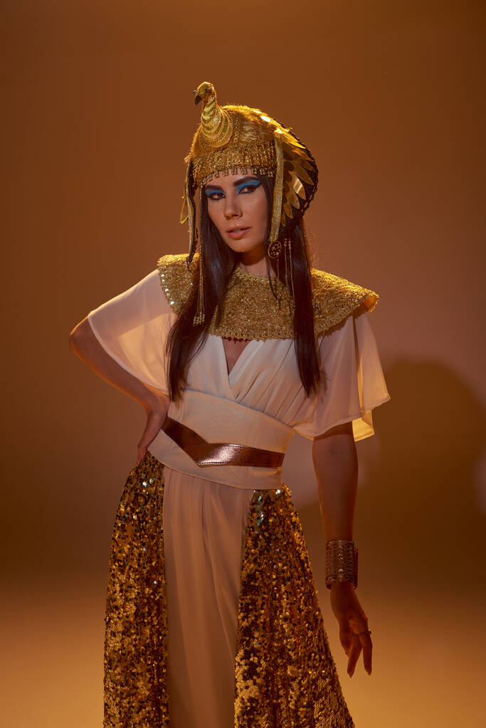 Stilvolles brünettes Model im ägyptischen Look und fettem Make-up posiert und steht auf braunem Hintergrund - Foto, Bild