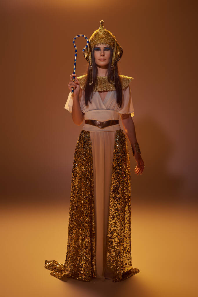 Volle Länge der stilvollen Frau in ägyptischer Kleidung, die Gauner hält und auf braunem Hintergrund steht - Foto, Bild