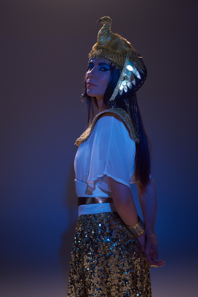 Elegante vrouw in gouden Egyptische look en hoofdtooi kijkend naar camera op bruin met blauw licht - Foto, afbeelding