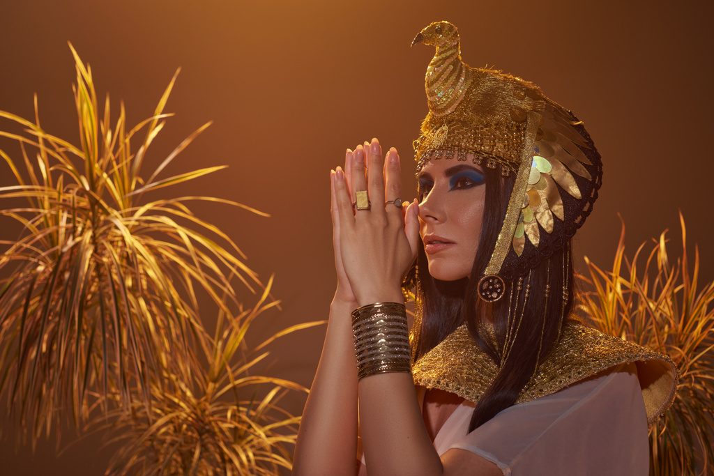 Brunette woman in egyptian look doing praying hands gesture near desert plants isolated on brown - Photo, Image