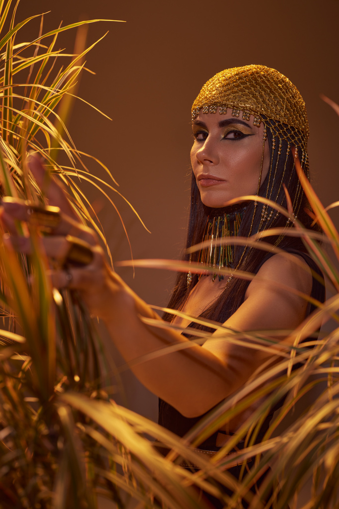 Stylish brunette woman in egyptian look touching plants and looking at camera isolated on brown - Photo, Image