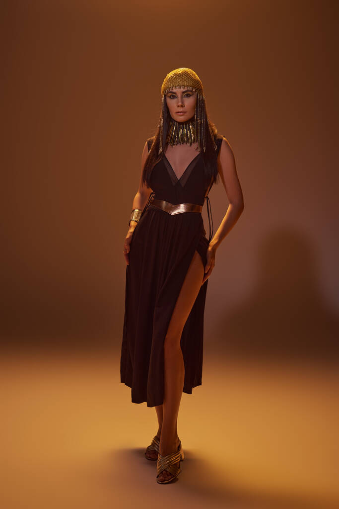 Volle Länge der eleganten Frau im ägyptischen Look und Kopfschmuck stehend und posierend auf braunem Hintergrund - Foto, Bild