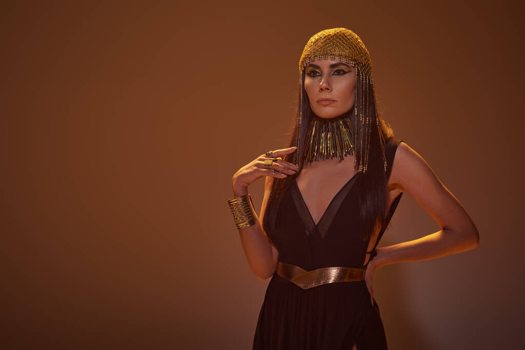 Trendy Frau in ägyptischem Look und Halskette hält Hand auf Hüfte, während sie auf braunem Hintergrund steht - Foto, Bild