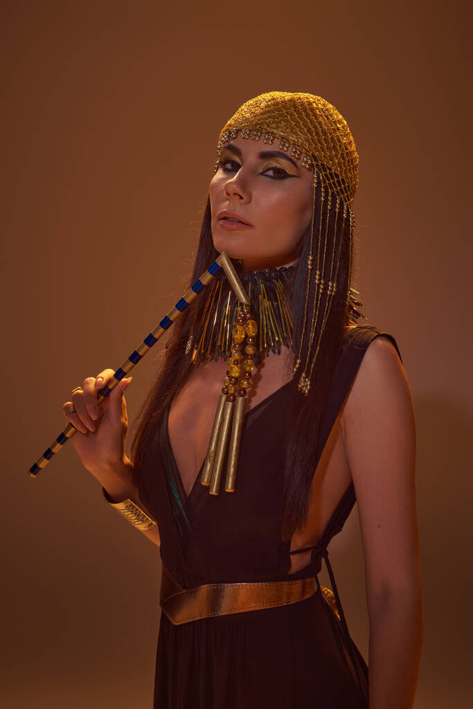 Elegante vrouw met make-up en Egyptische look houden vlegel en kijken naar de camera op bruine achtergrond - Foto, afbeelding