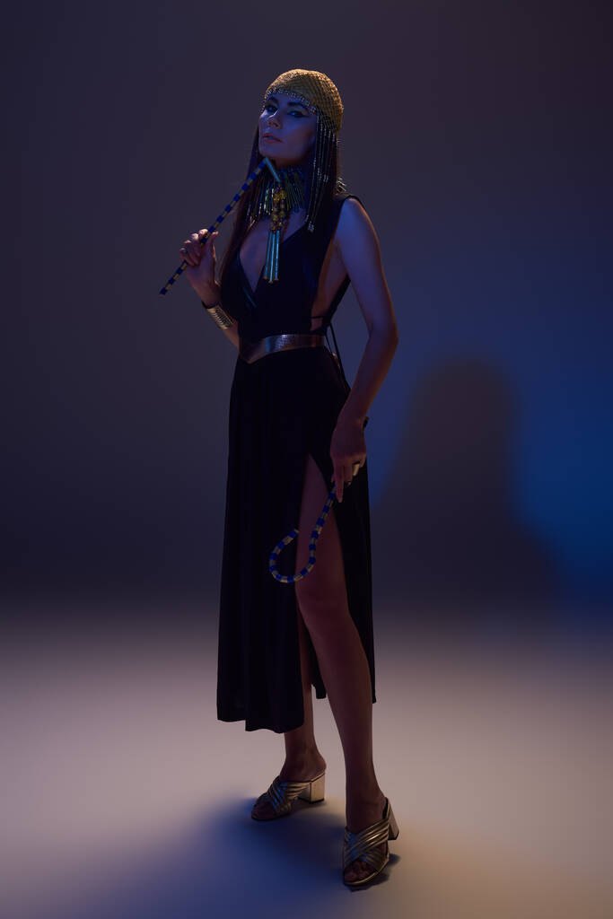 Stylish woman in egyptian look posing and holding crook and flail while standing on blue background - Photo, Image