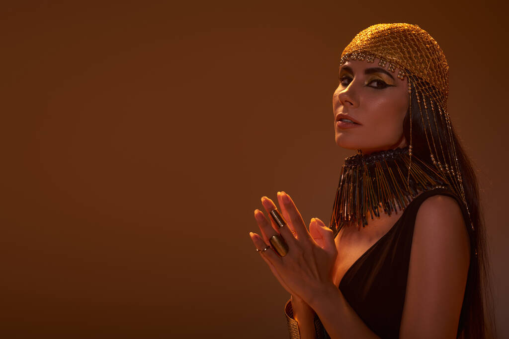 Mujer en tocado dorado y mirada egipcia mirando a la cámara aislada en marrón con luz - Foto, Imagen