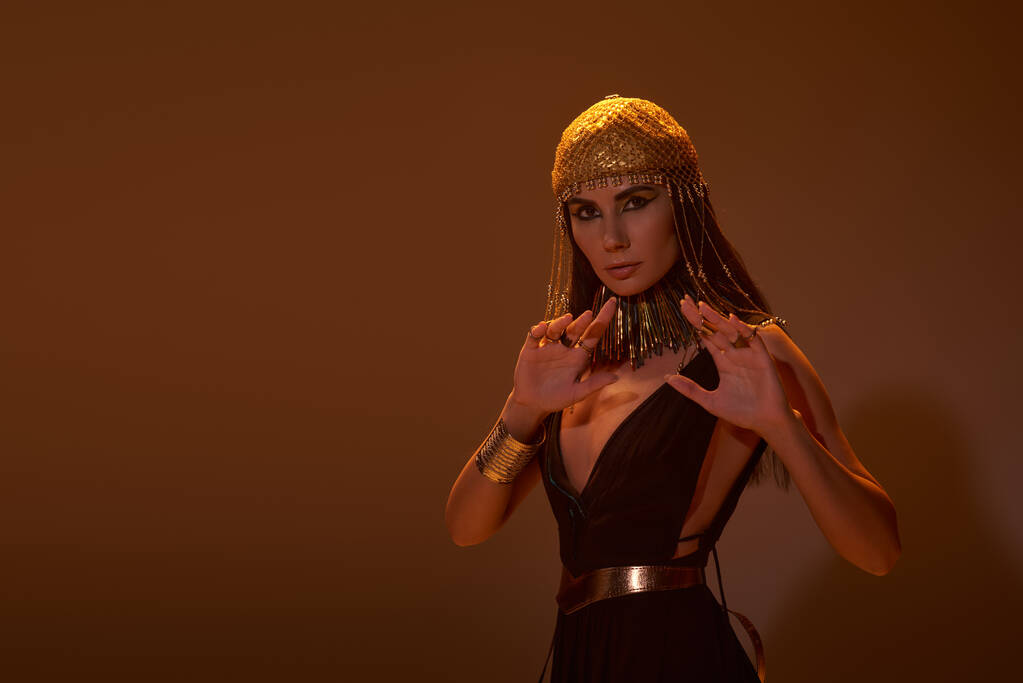 Kobieta w egipskiej sukience i nakryciu głowy pozowanie i patrząc na aparat na brązowym tle ze światłem - Zdjęcie, obraz
