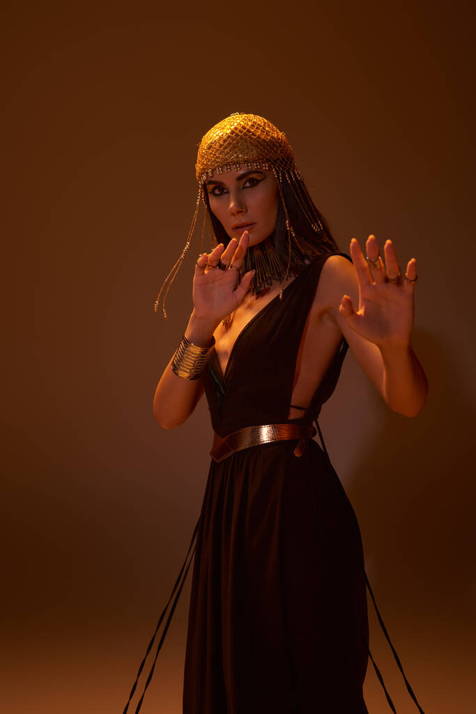 Brünette Frau im ägyptischen Kleid und Blick posiert und schaut in die Kamera isoliert auf braun - Foto, Bild