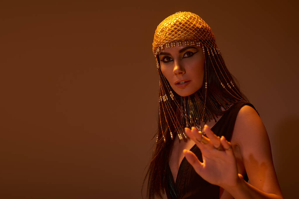 Женщина в египетском головном уборе и смотрит на камеру на коричневом фоне со светом - Фото, изображение