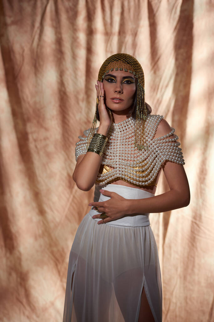 Elegante modello in look egiziano e top in perla toccante guancia e in piedi su sfondo astratto - Foto, immagini