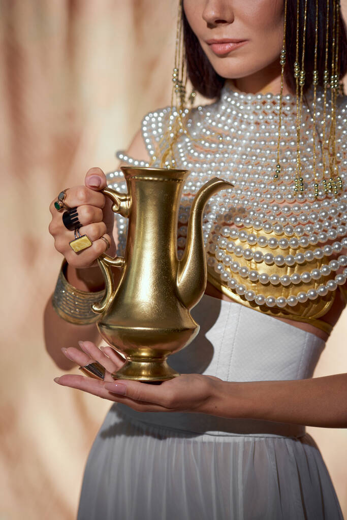 Vue recadrée de la femme élégante dans le regard égyptien tenant cruche d'or sur fond abstrait - Photo, image
