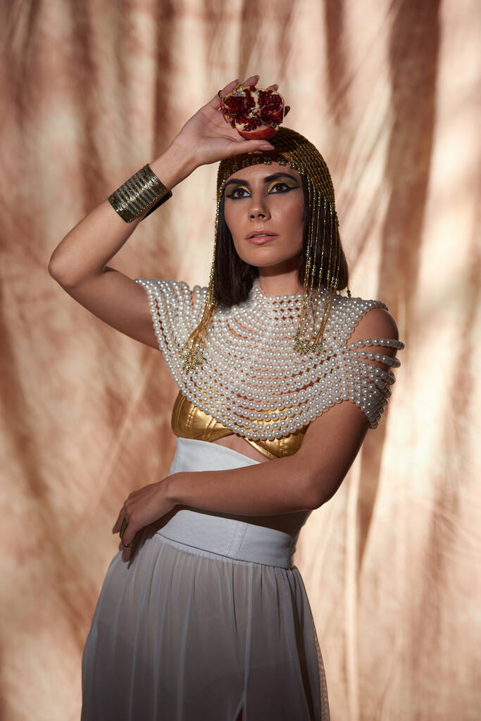 Стильна жінка в єгипетському вигляді і сміливий макіяж, що тримає вирізаний гранат на абстрактному фоні - Фото, зображення