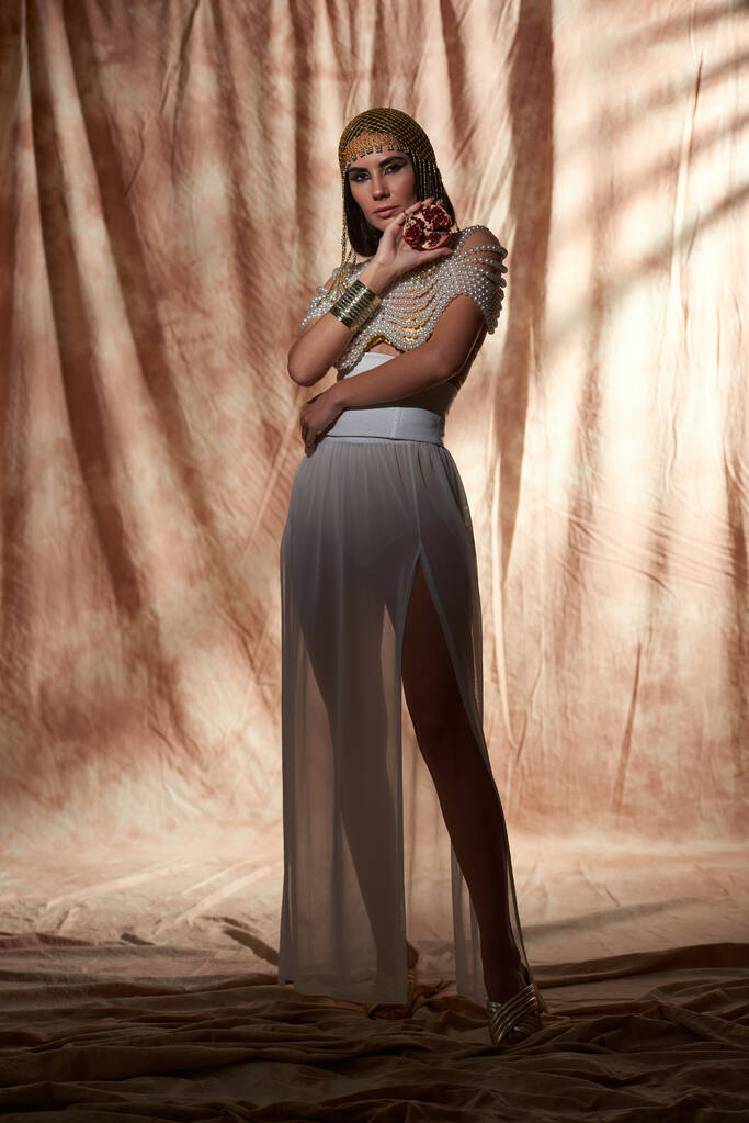 Plná délka ženy v egyptském oděvu a perleťové vrchní držení broušené granátové jablko na abstraktním pozadí - Fotografie, Obrázek