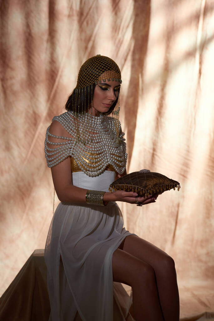 Женщина в египетском виде держит подушку с драгоценностями и драгоценными камнями, позируя на абстрактном фоне - Фото, изображение