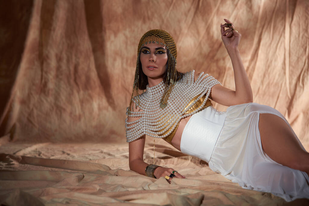 Элегантная женщина в египетском костюме и перламутровом топе лежит на абстрактном фоне с солнечным светом - Фото, изображение