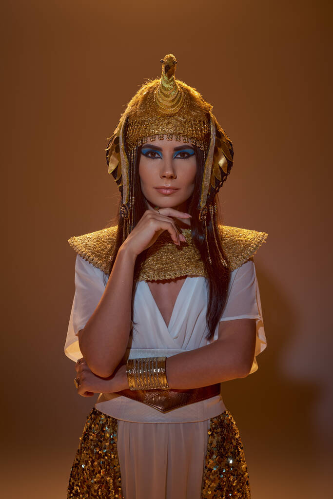 Stilvolle brünette Frau mit Make-up und ägyptischer Kleidung schaut in die Kamera auf braunem Hintergrund - Foto, Bild