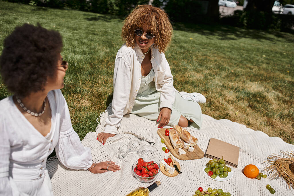 jong Afrikaans amerikaanse vriendinnen in witte jurken ontspannen op de zomer picknick in de buurt van fruit en wijn - Foto, afbeelding