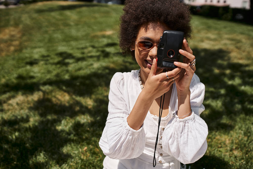 heureuse femme afro-américaine en lunettes de soleil prenant des photos sur caméra vintage dans le parc d'été, joie - Photo, image