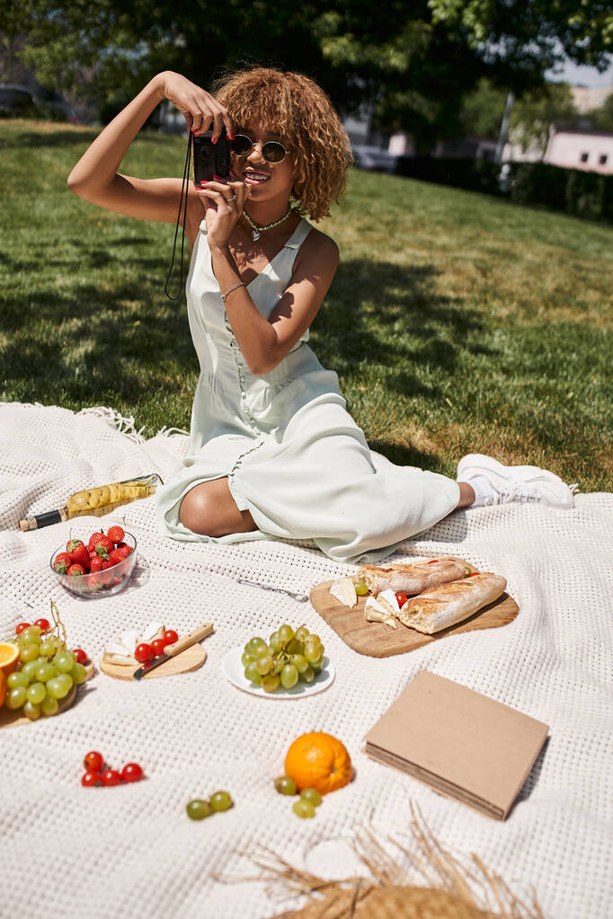 ピクニックで野菜や果物の近くのヴィンテージカメラで写真を撮る楽しいアフリカ系アメリカ人女性 - 写真・画像