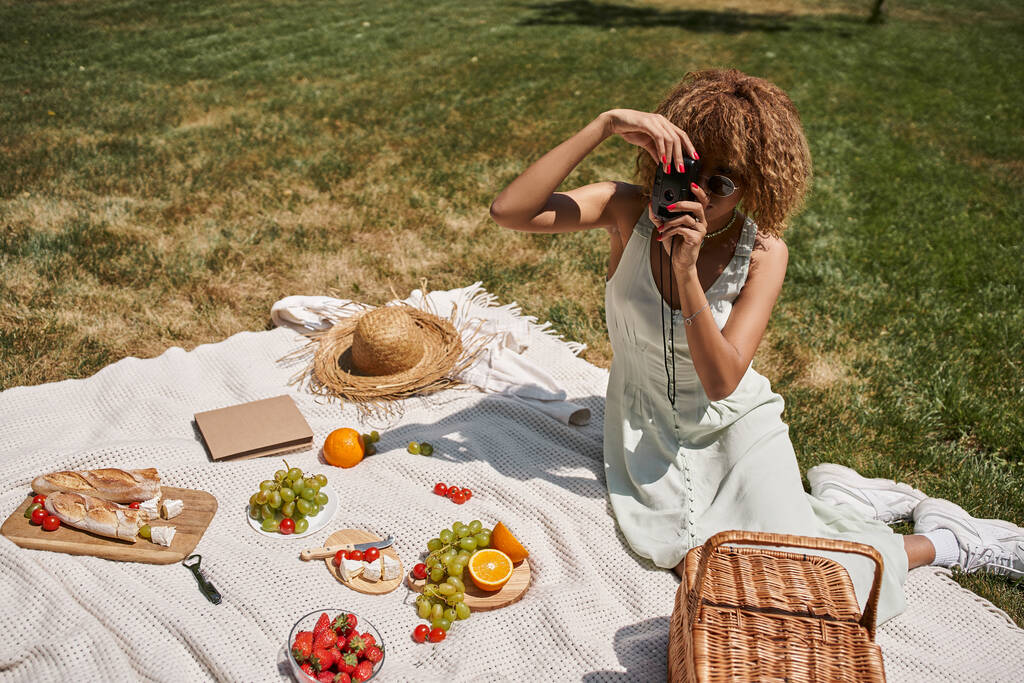 афроамериканська жінка робить фото на старовинній камері біля фруктів і овочів, літній пікнік - Фото, зображення