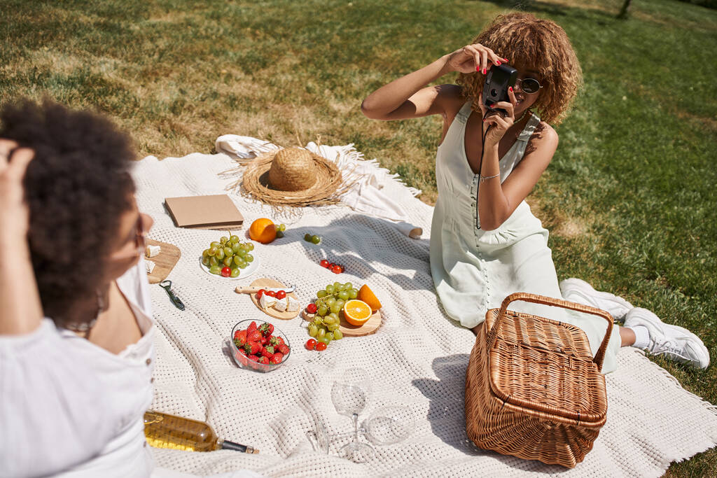 young african american woman taking photo of girlfriend on vintage camera, picnic in summer park - Photo, Image