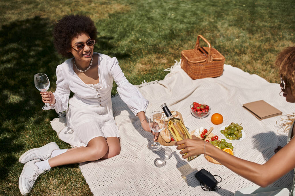 carefree african american girlfriends pouring wine near fruits and vegetables during picnic - Photo, Image