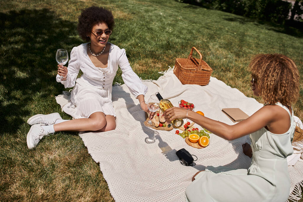 happy african american woman holding glasses near girlfriend pouring wine during summer picnic - Photo, Image