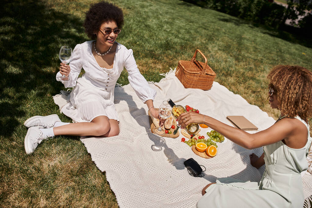 Afrika kökenli Amerikalı bir kadın kız arkadaşının yanında şarap bardakları, sebzeler ve meyveler, yaz pikniği - Fotoğraf, Görsel