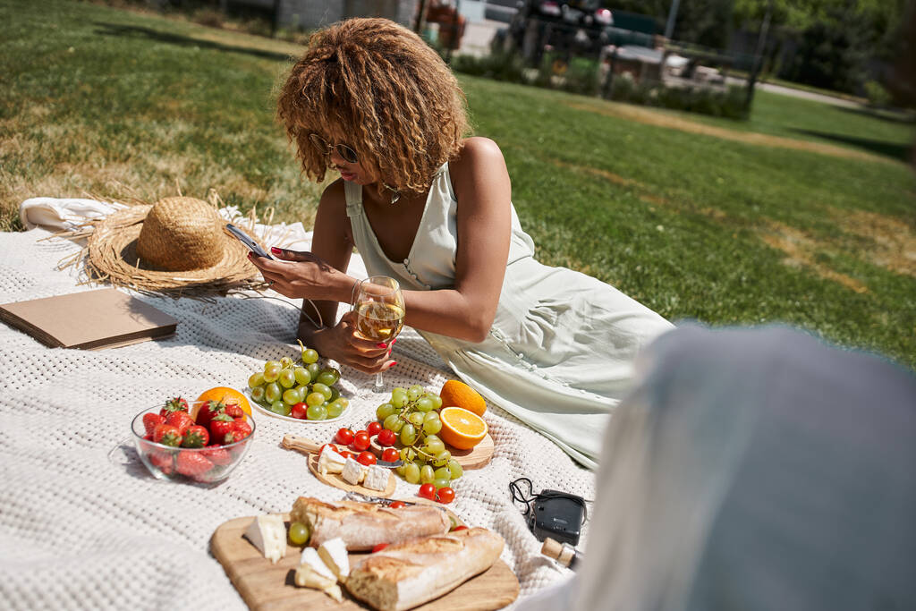 Αφροαμερικανή γυναίκα με γυαλί κρασιού χρησιμοποιώντας smartphone κοντά σε φρούτα και φίλη για πικνίκ στο πάρκο - Φωτογραφία, εικόνα
