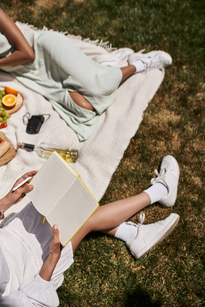afro-américaine avec ordinateur portable près de petite amie, fruits et bouteille de vin sur couverture dans le parc - Photo, image