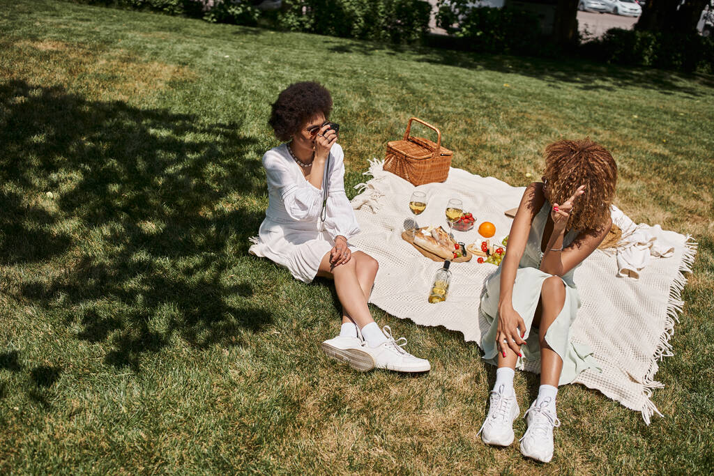 Африканская американка с винтажной камерой фотографирует подружку на лужайке во время летнего пикника - Фото, изображение