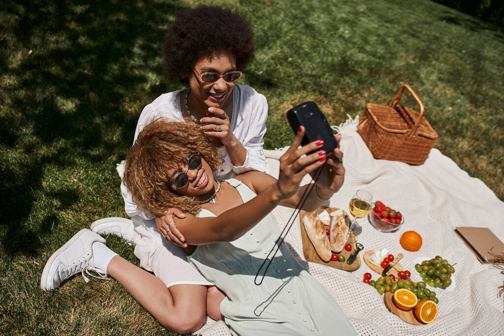 top view of joyful african american girlfriends taking selfie near food and wine, picnic in park - Photo, Image