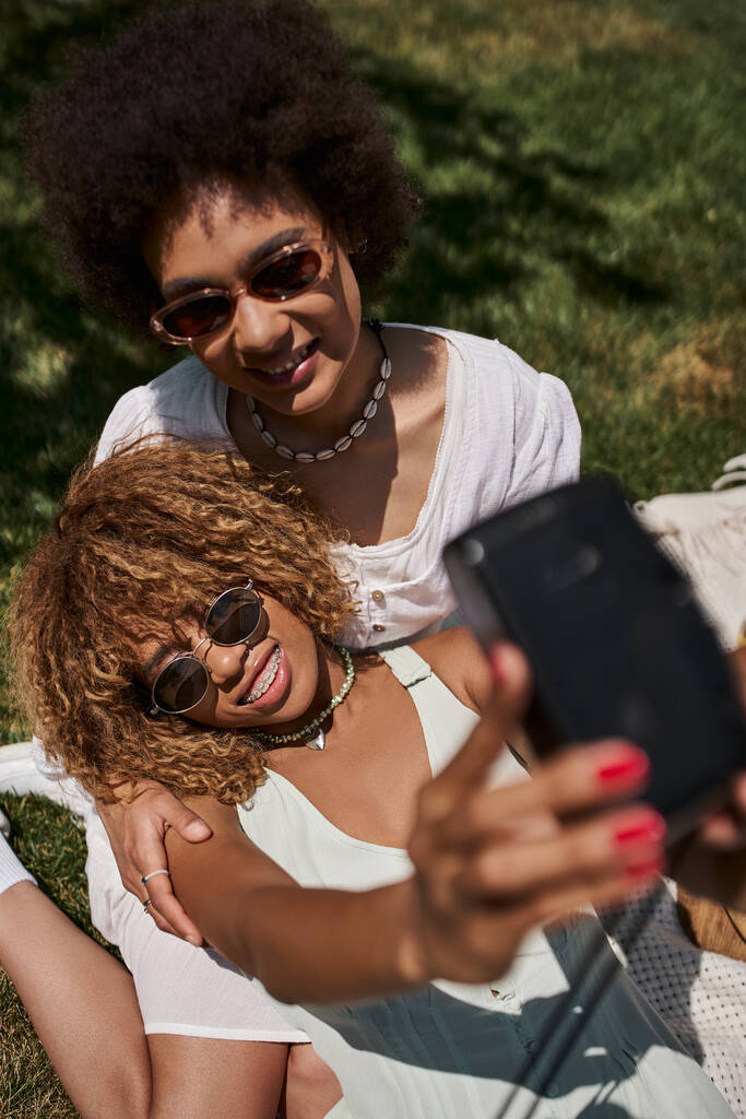 happy and stylish african american girlfriends in sunglasses taking selfie on vintage camera in park - Photo, Image